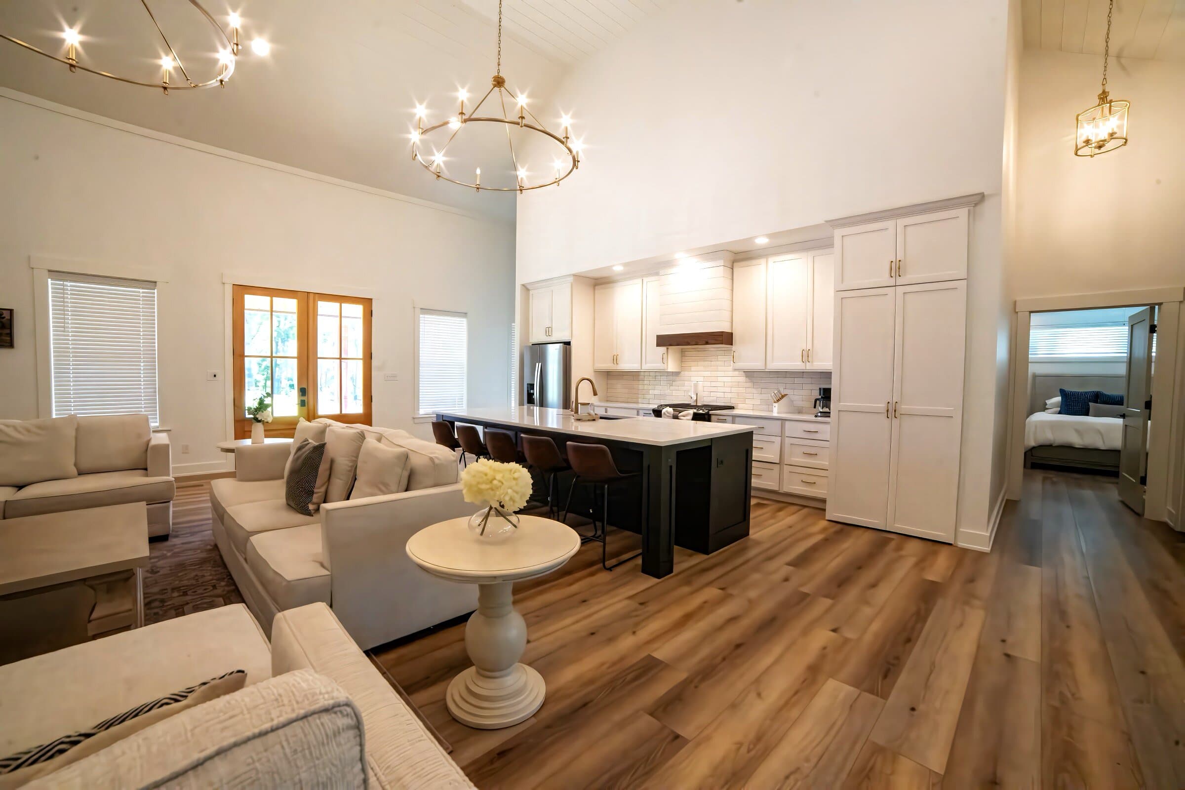 Vaulted great room with white kitchen, brass chandeliers, and cream sectional